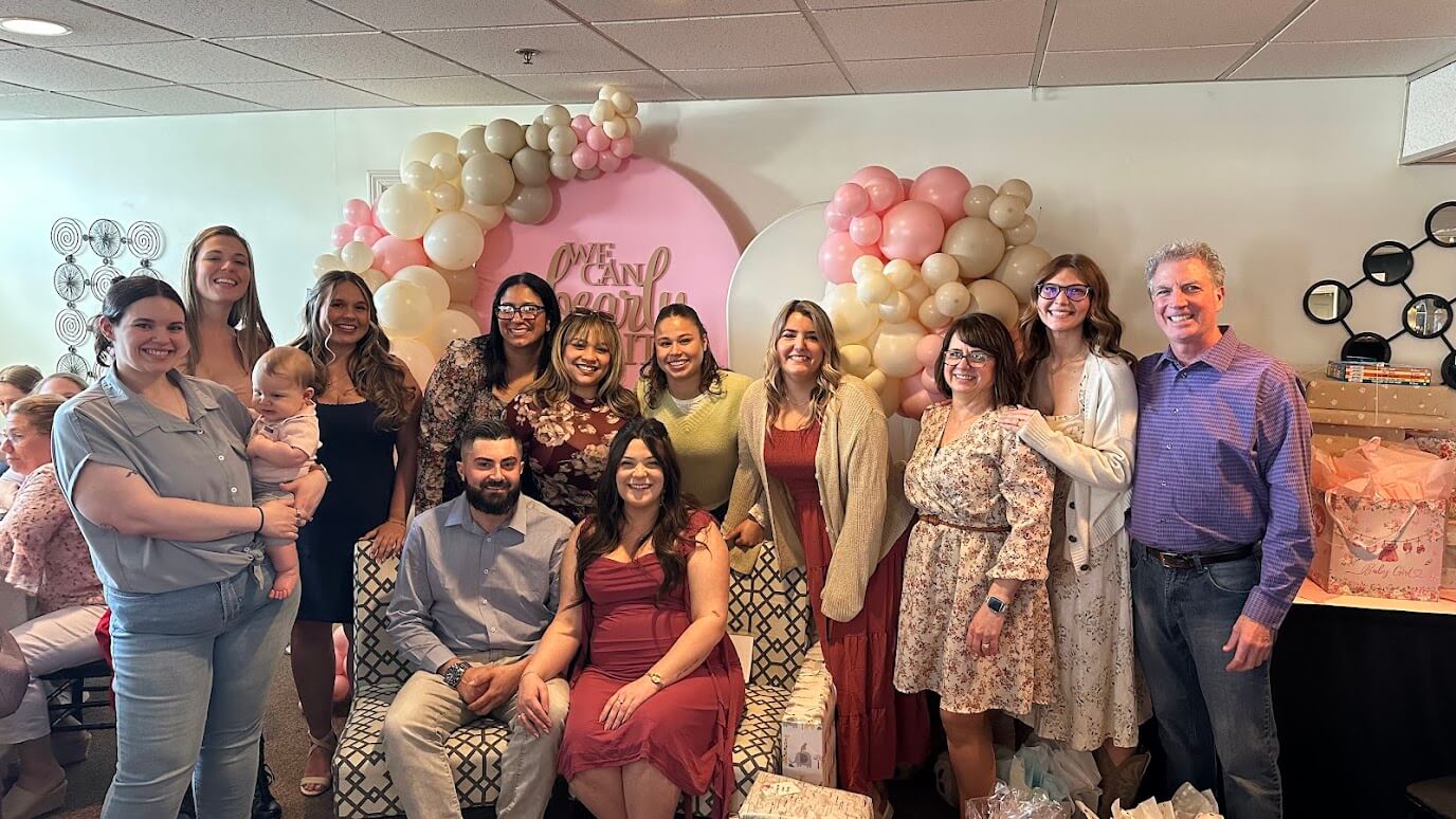 Dental team gathered at an office baby shower with a pink balloon arch in Lunenburg