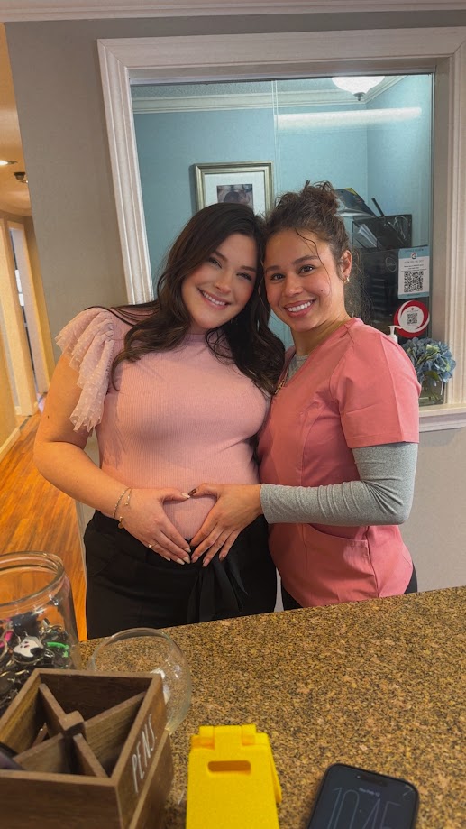 Two dental team members making a heart shape over a baby bump at the front desk in Lunenburg
