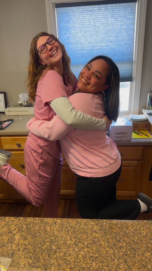 Two dental team members in pink scrubs laughing and hugging in the office in Lunenburg