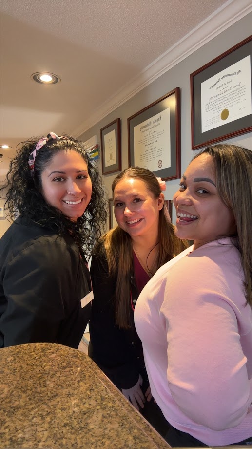 Three dental team members smiling together near the front desk and diploma wall in Lunenburg