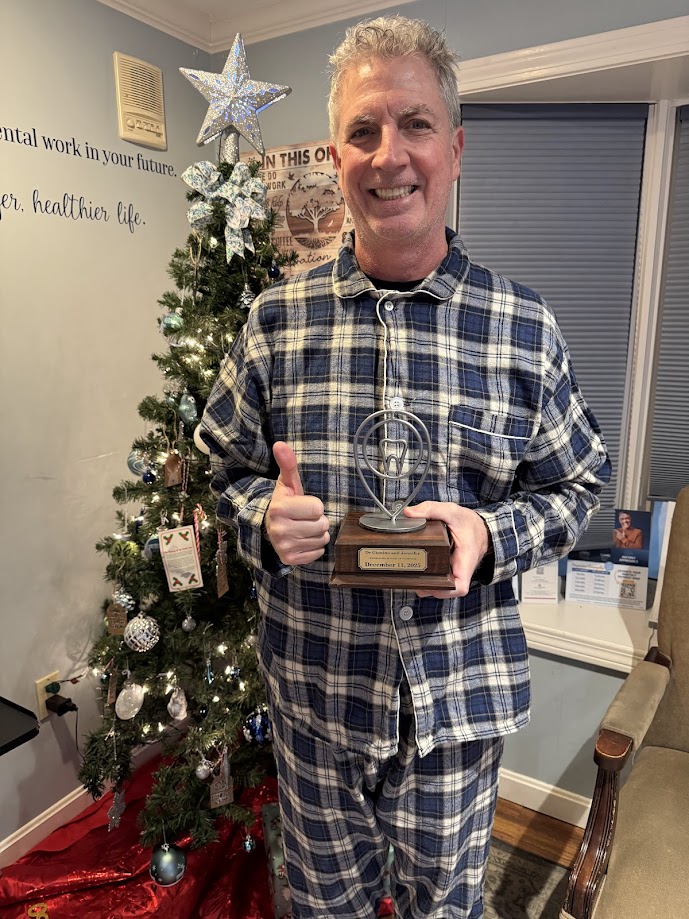 Dentist in holiday pajamas holding a teamwork award beside the office Christmas tree in Lunenburg